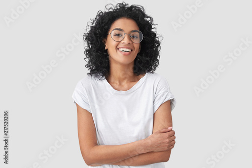 Fotografía Joyful African American student keeps hands crossed, laughs at good joke, wears casual clothes and round spectacles, isolated over white background