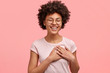 canvas print picture - Photo of friendly positive female smiles sincerely, has Afro hair, keeps both palms on heart, dressed in casual t shirt, isolated over pink wall, expresses good feelings and attitude to close person