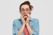 © WHstudio Leushin N - Embarrassed nervous female with freckled skin, keeps hands near mouth, looks nervously, being pleased by something pleasant, wears round glasses, isolated over white background. Emotions, reaction