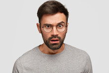 Headshot Of Handsome Unshaven Male Reacts On Something With Displeased Look, Has Surprised Expression, Stares At Camera, Dressed In Casual Clothes, Isolated Over White Background. Emotions Concept