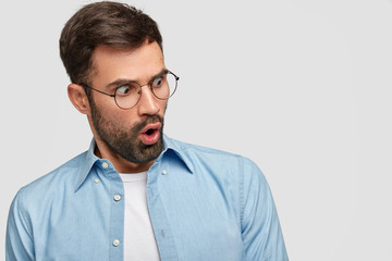 Scared surprised bearded man stares with stupefied expression, opens mouth widely, notices something awful, wears spectacles, poses against white background with copy space for your advertisement