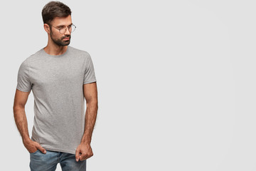 Fashionable young muscular male in grey casual t shirt and jeans, keeps hand in pocket, looks seriously aside, focused on something, poses against white blank wall on left for your advertisement