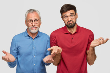 Wall Mural - Indoor shot of puzzled mature male and his adult son, shrugg shoulders, can`t make decision, being unshaven, stand closely to each other against white background. So what to do in this situation?