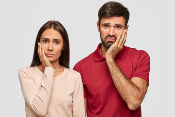 Wall Mural - Displeased young female and male keep hands on cheeks, look with gloomy expressions, feel bored while watch not interesting film, dressed casually, spend time at home. Emotions and people concept