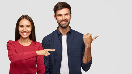 Wall Mural - Cheerful brunette young female in red sweater with healthy skin, positive bearded guy stand together in studio, point aside at blank space, show place for your advertisement or promotional text