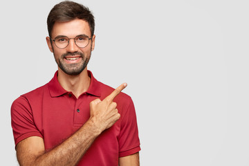 Wall Mural - Happy smiling European male with bristle, points with index finger aside, invites you to have dinner in restaurant situated near, dressed in casual red t shirt, spectacles isolated on white background