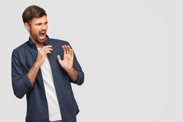 Wall Mural - Desperate irritated young bearded man with negative facial expression, says fie, pushes hands, sees something disgusting and unpleasant, stands agaist white background with copy space on right