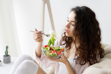 Wall Mural - Young girl woman female wear pajams sleepwear, white top and stockings sitting on sofa and eating getting breakfast green healthy salad in interior light room. Morning route concept