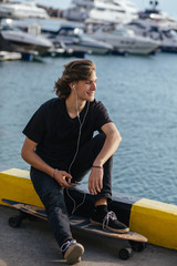 Wall Mural - Young man using a cellphone while sitting at longboard on pier witth yacht