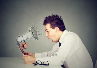 man sitting at table working on computer screaming with a megaphone poking out from a laptop screen