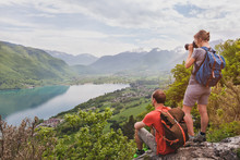 Travel And Tourism, Couple Of Travelers With Backpacks Enjoying Panoramic View Of Lake, Hikers Relaxing  On Top Of Mountain