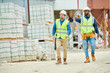 © Seventyfour - Two young men in hardhats and waistcoats pointing at distance while walking near stacks of building materials during inspection on construction site