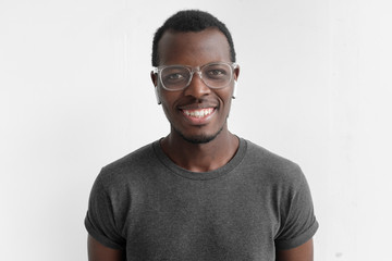 Wall Mural - Horizontal shot of young dark-skinned man with positive smile on face, isolated on gray background, wearing plastic glasses, being in high spirit because of personal success, dressed in casual T-shirt