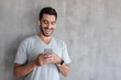 © Damir Khabirov - Indoor daylight picture of handsome man wearing gray casual t-shirt, laughing happily being amused by content on screen of smartphone he is holding in both hands, standing against textured wall