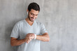 © Damir Khabirov - Young smiling man wearing t shirt and wireless headphones, holding smart watches with touch screen, standing against gray textured wall with copy space