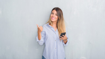 Sticker - Young adult woman over grey grunge wall looking at smartphone texting a message happy with big smile doing ok sign, thumb up with fingers, excellent sign