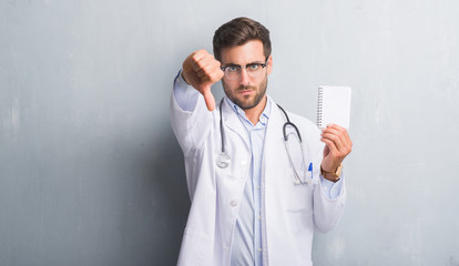 Handsome young doctor man over grey grunge wall holding blank notebook with angry face, negative sign showing dislike with thumbs down, rejection concept