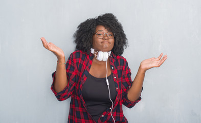 Poster - Young african american woman over grey grunge wall wearing headphones clueless and confused expression with arms and hands raised. Doubt concept.