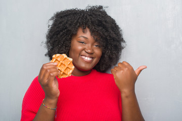 Poster - Young african american woman over grey grunge wall eating belgium waffle pointing and showing with thumb up to the side with happy face smiling