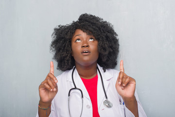 Poster - Young african american doctor woman over grey grunge wall amazed and surprised looking up and pointing with fingers and raised arms.