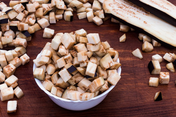 Eggplant, cut into small cubes, laid in a plate and scattered on the table, two longitudinal slices of eggplant with a blue peel on a wooden background.