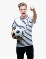 Sticker - Young handsome blond man holding soccer ball annoyed and frustrated shouting with anger, crazy and yelling with raised hand, anger concept