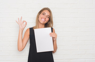 Beautiful young woman over white brick wall holding blank paper sheet doing ok sign with fingers, excellent symbol