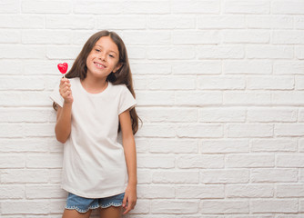 Sticker - Young hispanic kid over white brick wall eating red heart lollipop candy with a happy face standing and smiling with a confident smile showing teeth