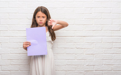 Poster - Young hispanic kid over white brick wall holding pink paper sheet with angry face, negative sign showing dislike with thumbs down, rejection concept