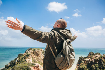 Wall Mural - View from the back. A young man tourist enjoys the beautiful views of the Atlantic Ocean and the landscape off the coast in Portugal and raises his hands upward showing how pleased he is.