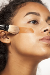 Wall Mural - portrait of a young dark-skinned woman applying liquid makeup base with brush on her face on a white background