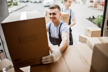 A Young Handsome Smiling Mover Wearing Uniform Is Reaching For The Box While Unloading The Van Full Of Boxes. House Move, Mover Service.
