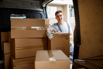 Wall Mural - A young handsome smiling worker wearing uniform is standing next to the van full of boxes. House move, mover service.