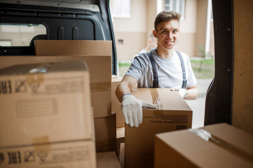 Wall Mural - A young handsome smiling worker wearing uniform is standing next to the van full of boxes. House move, mover service.