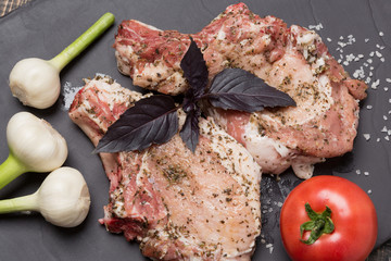 two pieces of steak with spices on black slate, young garlic and tomatoes, raw meat and basil leaves, large sea salt, concept of nutrition, close-up