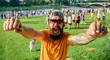 Urban event celebration. Hipster in cap happy celebrate event picnic fest or festival. Cheerful fan at summer fest. Man bearded hipster in front of crowd people raise fists green riverside background