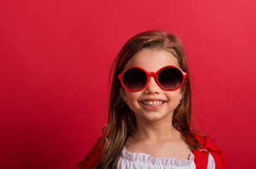 Wall Mural - Portrait of a small girl with red sunglasses in studio on a red background.