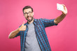 I love selfie! Handsome young man in shirt holding camera and making selfie and smiling while standing against pink background. Listening music with headphones.