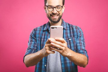 Wall Mural - Always in touch. Smiling young man holding smart phone and looking at it. Portrait of a happy man using mobile phone isolated over pink background.