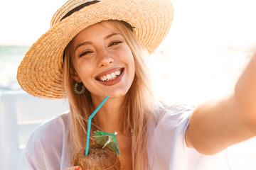 Sticker - Photo of happy young woman 20s in summer straw hat laughing, and drinking cocktail while taking selfie on beach during sunrise