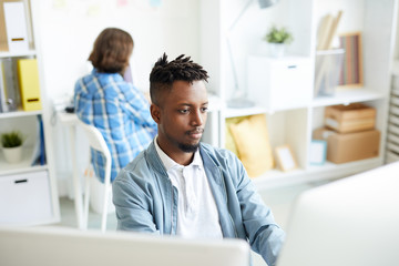 Wall Mural - young african-american man sitting by workplace in front of two desktop computers wotking with datab