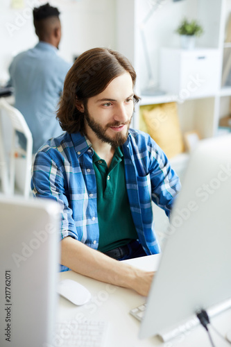 Cuadros en Lienzo Young bearded man in casualwear sitting in front of computer monitor and carryin