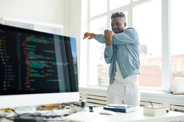 Sticker - young it-manager in casualwear standing between window and his desk with computer and doing exercise