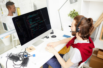 Wall Mural - young it-manager in headset sitting in front of computer monitor, talking to clients and working wit