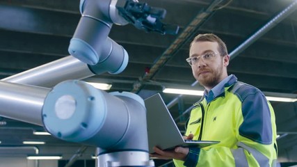 Fototapete - Low Angle Shot At the Factory: Automation Engineer Uses Laptop for Programming Robotic Arm. New Era in Automatic Manufacturing Industry.
