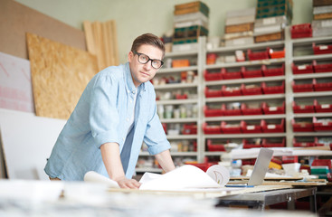 Young handcraft master leaning over workplace with sketch and looking at you during working process