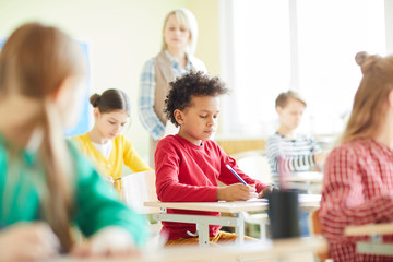 Serious concentrated African-American boy with Afro hairstyle sitting at desk and doing quiz at school