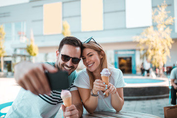 Wall Mural - Young man with sunglasses is taking selfie with his girlfriend while they having ice-cream outdoors