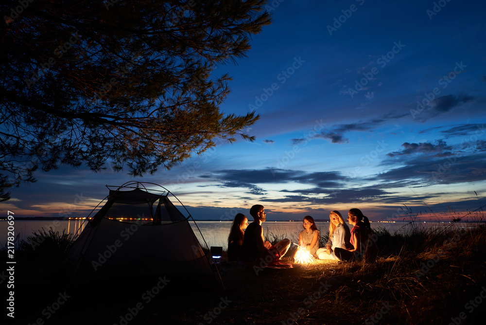 Night summer camping on lake shore. Group of five young tourists sitting on the beach around campfire near tent under beautiful blue evening sky. Tourism, friendship and beauty of nature concept. - obrazy, fototapety, plakaty 