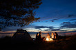 Night summer camping on lake shore. Group of five young tourists sitting on the beach around campfire near tent under beautiful blue evening sky. Tourism, friendship and beauty of nature concept.